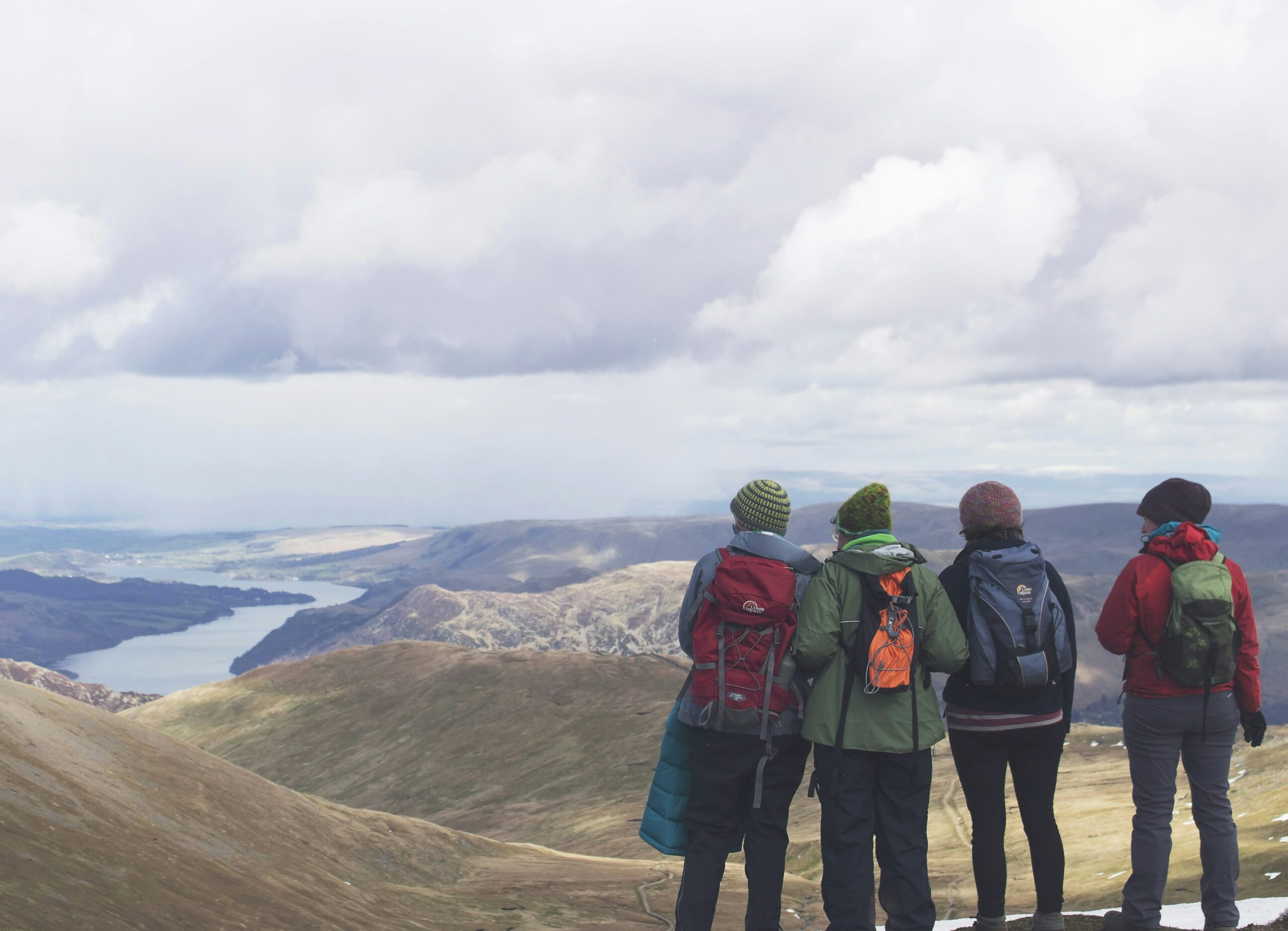 Group of four hikers with backpacks standing on a mountain summit, looking out over a scenic valley and lake below under a cloudy sky.