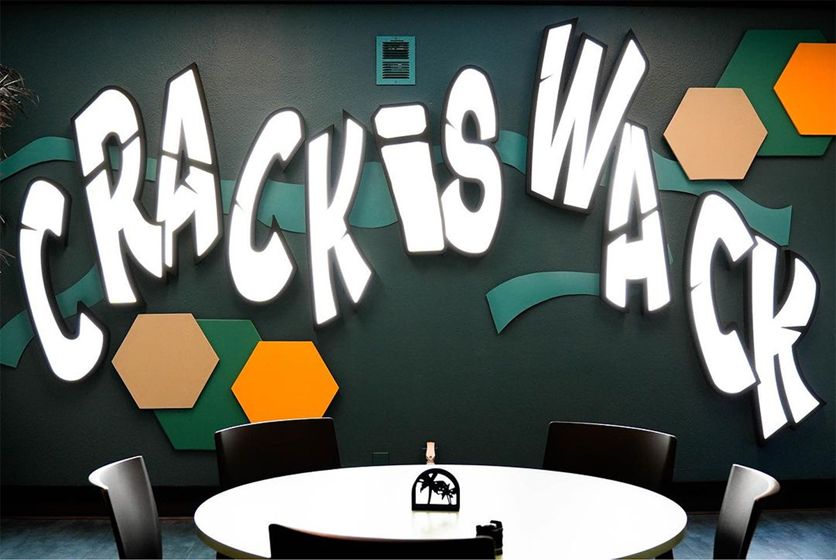 Bold mural with the phrase 'Crack is Wack' in large white letters on a dark wall, behind a round table and chairs in a day treatment facility.