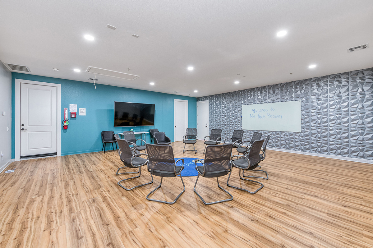Group therapy room at a residential treatment center with circular chair arrangement, wood flooring, textured wall design, and a flat-screen TV.